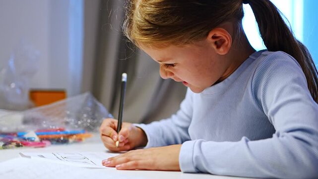 First grade girl writes with pencil while studying at home. Focused child develops handwriting and learning skills with selective focus and shallow depth of field