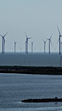 Vertical panning shot of offshore wind turbines rising from IJsselmeer lake beyond dike and rock breakwater near Breezanddijk on Afsluitdijk in Netherlands
