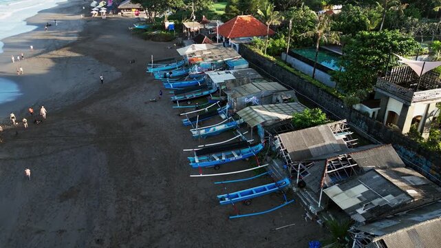 Traditional wooden jukung fishing boats lined up on sandy shore with local coastal shacks and tropical resort in bright morning sunlight Bali orbit aerial drone footage