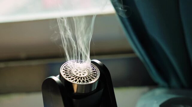 slow motion shot of white smoke or vapor gently rising and swirling from a small, sleek black diffuser with a metallic patterned grill. The portable air freshener or mini humidifier is operat