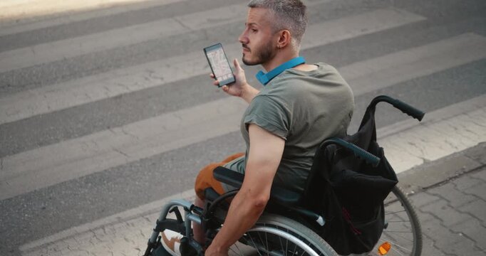 Young man in a wheelchair uses GPS navigation on a smartphone while moving through the city, checking the map and following a route during everyday independent mobility.
