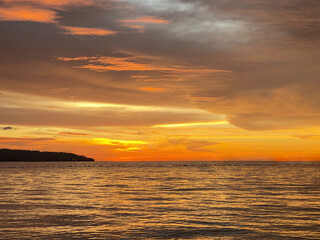 Fototapeta premium Dramatic golden sunset over calm ocean water with colorful clouds and a peaceful coastal horizon, creating a beautiful tropical seascape.