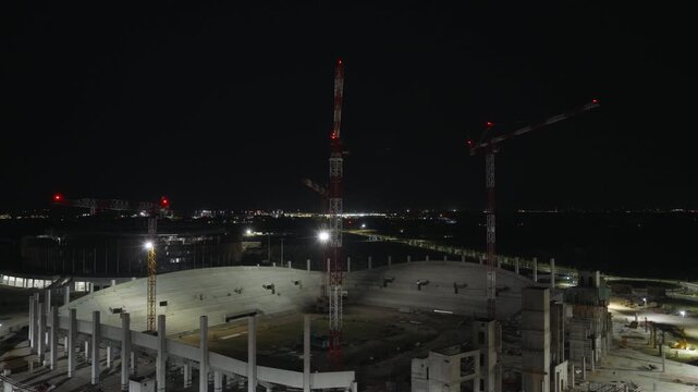 Multiple tall red cranes rise above oval concrete stadium framework at Venezia FC construction site near Venice, distant city lights glow beneath dark starless night sky. Aerial drone orbit shot