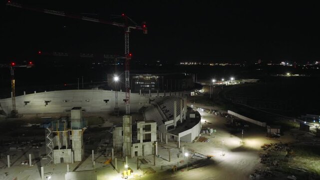 Curved concrete stands take shape at new Venezia FC stadium construction site in Venice, tall red crane towers above brightly illuminated structure beneath dark night sky.Aerial drone approaching shot
