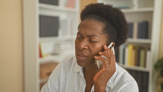 Middle aged african american woman holding smartphone to ear, frowning with hand at jaw, seated near bookshelf in building; concerned.