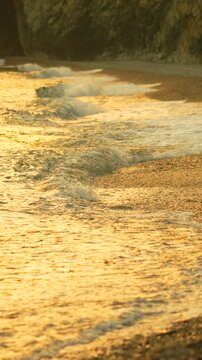 Golden ocean tide rolls gently onto pebble shore at dusk beneath steep coastal cliffs. High angle vertical static shot.