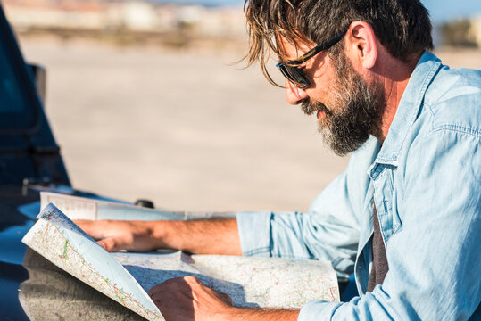 Bearded man in sunglasses leans on a car hood studying a folded road map, planning a coastal road trip in bright daylight, concentrating on navigation and route details now.