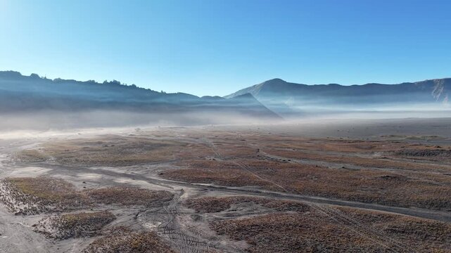 Mist curls over rugged, barren earth. Dusty trails wind through arid, sunlit plains near Bromo