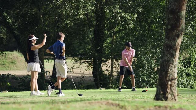 Three teenage friends playing a round of golf on a lush green course. One player tees off with a driver as his friends watch and cheer