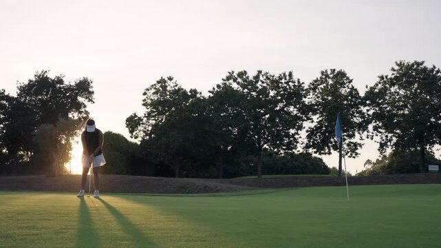 Young female golfer concentrating and hitting a golf ball with a putter on a green course. Beautiful sunset light creates long shadows