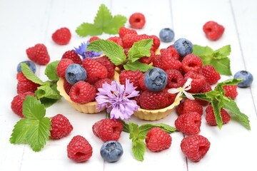 Fresh organic raspberries with tartlets, fresh green mint leaves, and purple cornflowers on white wooden background, sweet summer berry dessert © Andrii