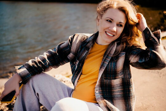 Smiling woman in plaid shirt enjoys autumn travel sitting by water. Cheerful female tourist rests near river looking away. Solitude in pleasure, mental wellbeing, peaceful outdoor nature retreat.