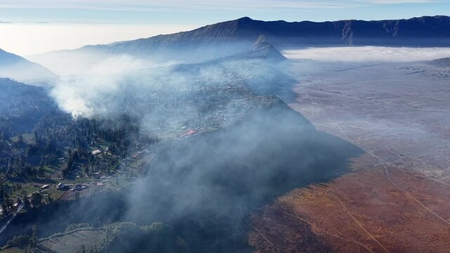 Smoke rises over mountainous terrain at Cemorolawang, drifting through valleys. Fog blankets distant peaks