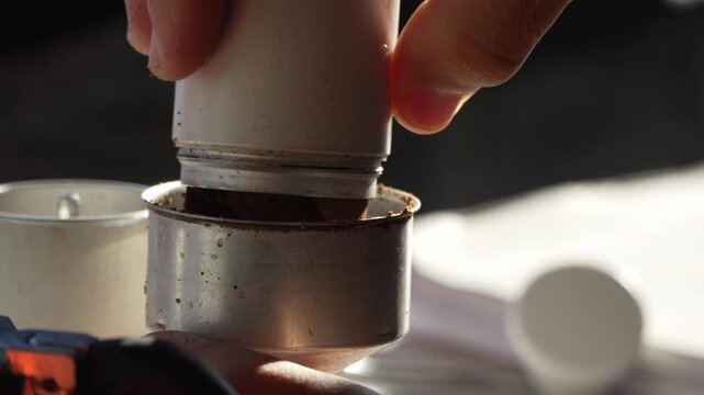 Fresh coffee grounds poured into a moka pot filter basket outdoors in the sunlight during morning camping preparation. Close-up shot.