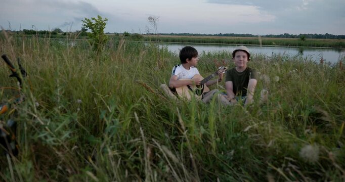 Two boys play acoustic guitar playing music enjoying nature. Teenagers spending time together. 4K 10-bit