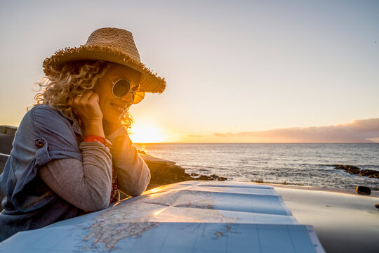 Woman wearing straw hat and sunglasses studies a map on a car roof at sunset by the sea, smiling thoughtfully during a coastal road trip and planning a relaxed travel adventure.