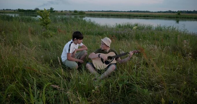 Two boys play acoustic guitar playing music enjoying nature. Teenagers spending time together. 4K 10-bit