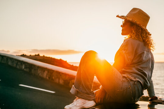 Woman in straw hat and sunglasses sits on a seaside ledge at golden hour, watching sunset over the ocean, feeling peaceful, enjoying warm light and quiet coastal evening solitude.