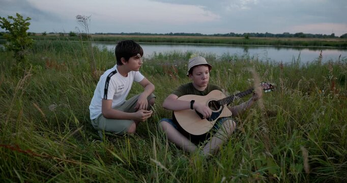 Two boys play acoustic guitar playing music enjoying nature. Teenagers spending time together. 4K 10-bit