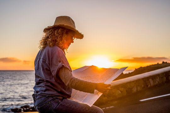 Woman wearing a straw hat reads a paper map at sunset by the ocean, planning a coastal route while seated on a low stone wall in warm golden light and feeling reflective and adventure