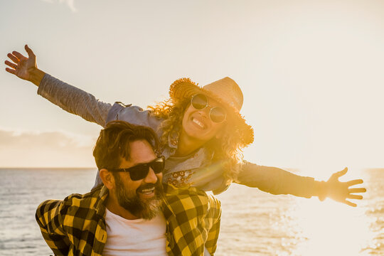 Joyful couple at the beach at sunset, woman on man's back, both wearing sunglasses and smiling as golden light reflects on the sea, conveying freedom, warmth and playful romance.