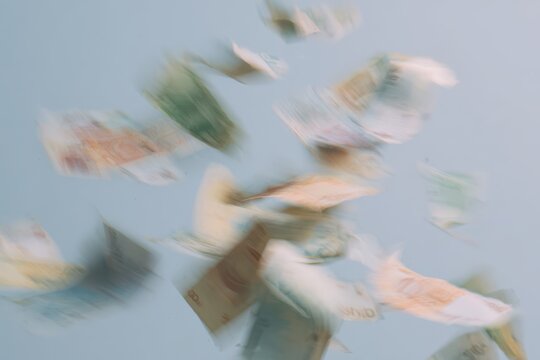 Money falls in a slow motion blur with different currencies in shades of green, blue, and brown against a light background
