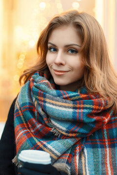 Young beautiful girl in stylish knitted scarf and black coat drinking coffee on the background of shop windows