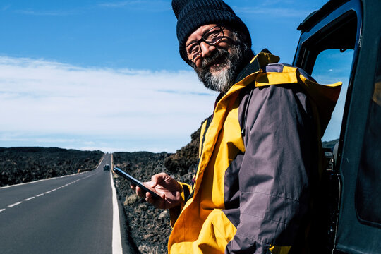 Smiling bearded man in yellow jacket leans against a car on an empty coastal road, checking his phone during a solo road trip under clear blue sky, relaxed and adventurous mood.