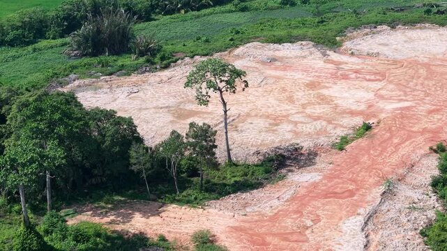 Aerial view shows deforested land with exposed red soil. Green trees stand near cleared ground