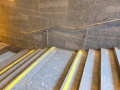 View of granite stairs with yellow tactile safety strips and metal handrail along stone wall.