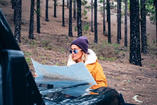 Woman in warm orange jacket and beanie studies a paper map on a car hood while parked in a pine forest, planning a road trip route and enjoying quiet outdoor exploration.