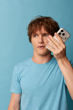 Surprised young man in a blue t-shirt holds a smartphone beside his cheek and glances sideways, startled by unexpected audio. Studio shot on blue background. Concept of privacy and security.