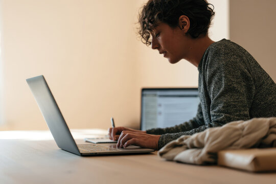 A young woman, focused on her laptop screen, writes notes, depicting a productive workspace filled with soft light, ideal for concentration and creativity.