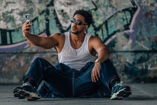 Confident young man taking a selfie with a smartphone while sitting on the ground in front of a colorful graffiti wall, cool urban street latin lifestyle portrait with a relaxed