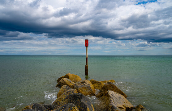 Red navigation marker and sea defence rocks at Horsey Gap, Norfolk, beneath dramatic storm clouds over the green-blue North Sea.