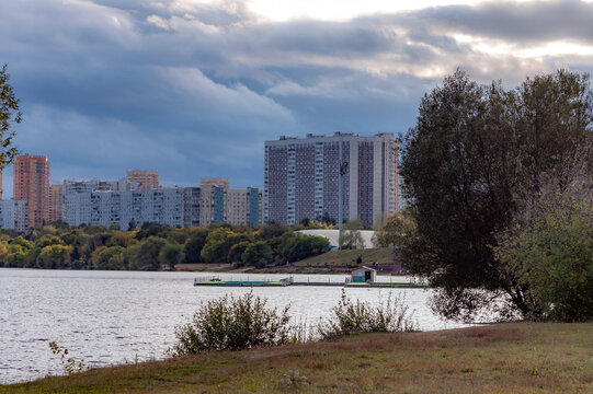 Scenic evening view of the river floodplain with dramatic clouds, sun rays shimmering on water, and residential apartment buildings, Strogino, Moscow, Russia.