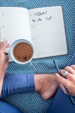 Holding a coffee while writing in a weekly planner with 'Today to do list' handwritten, relaxed planning on a patterned blanket with pen, legs crossed and a quiet routine.