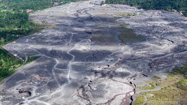 High angle view of heavy trucks and excavators harvesting rich volcanic sand deposits from a Semeru mudflow path.