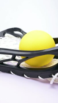 Bright yellow lacrosse ball resting inside a black lacrosse stick head with white woven netting rotating on white at vertical composition