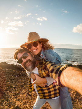 Couple taking a joyful seaside selfie at sunset, woman on man's back wearing a straw hat and sunglasses, both smiling warmly while rocky shore and ocean reach the horizon.