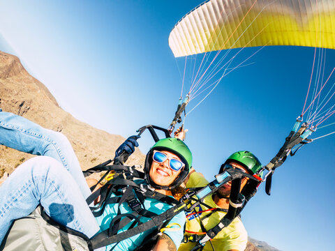 Smiling tandem paragliding couple flying over desert mountains with colorful canopy, wearing helmets and sunglasses, enjoying adrenaline adventure, freedom, travel, outdoors thrill.