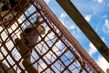 Young child climbing a rope net on a playground, viewed from below against the blue sky, showing active childhood recreation, adventure, movement and outdoor exploration © JairoMZ