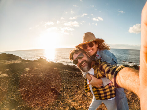 Couple on a playful piggyback on rocky coastline at sunset, wearing sunglasses and a hat while taking a selfie celebrating travel, romance, adventure and coastal freedom.