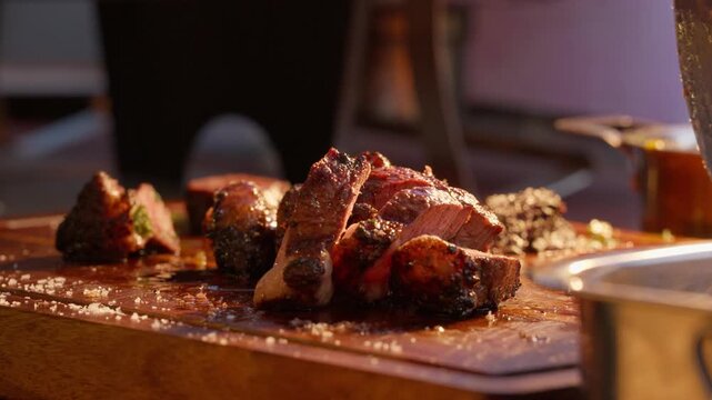 Grilled meat is carved into pieces with a large knife on a salted wooden cutting board during catering service at an outdoor event.