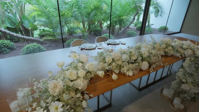 A long garland of white roses and daisies runs down a wooden banquet table set for dinner in a modern venue with glass walls facing a garden.