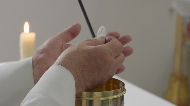 Close up of a priest holding a communion wafer above a gold ciborium at the altar during Catholic mass, with a lit candle behind.