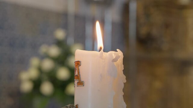 A thick altar candle with melted wax burns in a Catholic church beside white flowers, with blue azulejo tiles on the wall behind.