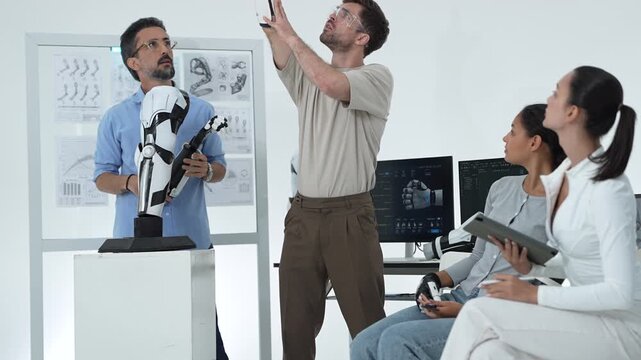 Engineer points a robotic forearm across the lab like a pointer, team reacts and laughs as they test the bionic hand prototype together