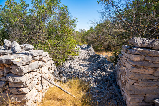 Rocky pathway between stone walls surrounded by green vegetation under a clear blue sky in a natural landscape setting next to Gornje Selo, Solta island, Croatia