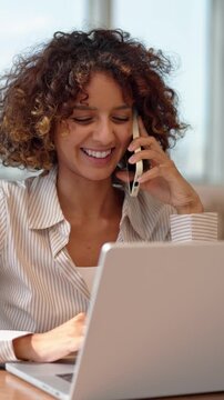 Vertical video of curly haired multiracial business woman in 30s talking on mobile phone call, sitting at laptop computer inside modern coworking office, discussing project with client, medium shot.